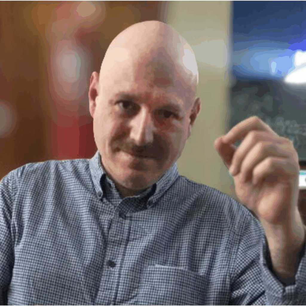 A bald man with a mustache, wearing a blue checkered shirt, sits indoors and gestures with his right hand. The background is blurred, showing warm colors and soft lighting.