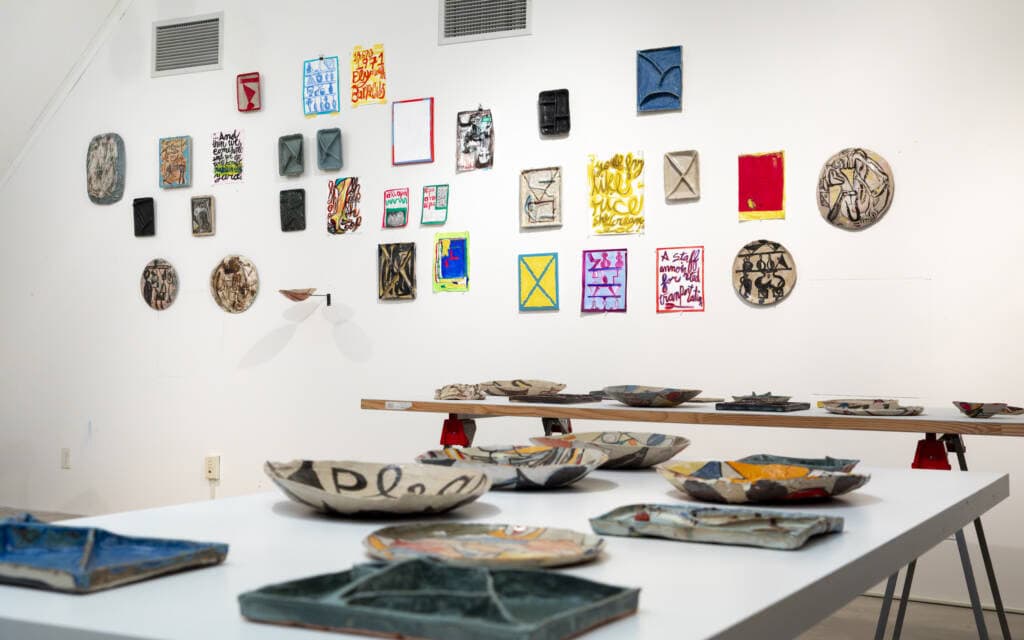 A gallery space with colorful abstract paintings and ceramic plates displayed on a white wall; ceramic dishes with various designs are arranged on white tables in the foreground.