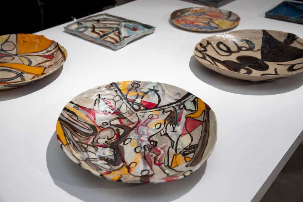 Several ceramic plates with colorful, abstract designs are displayed on a white table. The patterns feature bold lines, geometric shapes, and a mix of yellow, red, black, and blue tones.