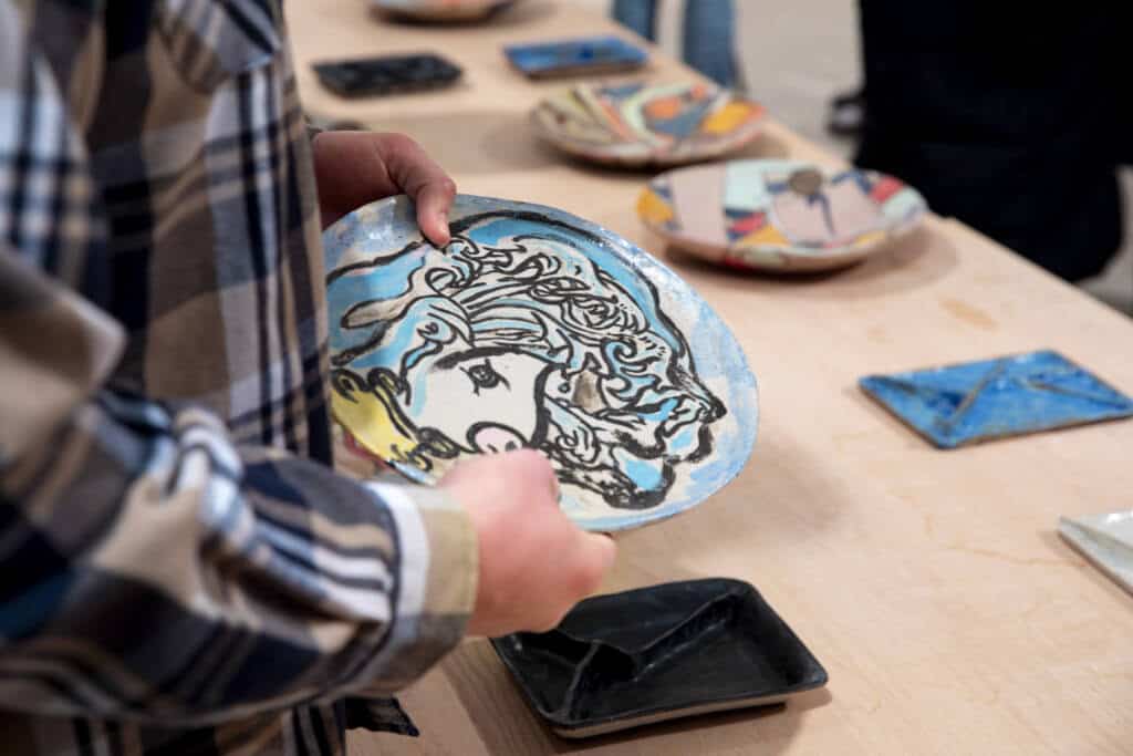 A person wearing a plaid shirt holds a hand-painted ceramic plate with an abstract face design. Other colorful plates and ceramics are displayed on a light wooden table in the background.