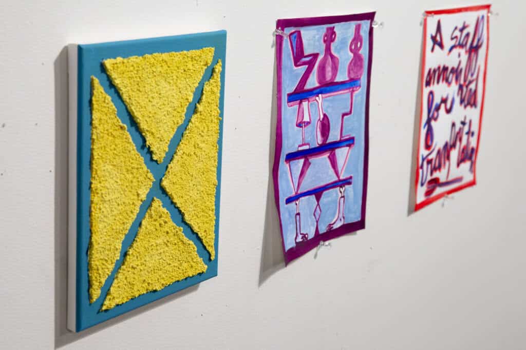 Three colorful art pieces hang on a white wall: a textured yellow and blue geometric panel, a pink and blue abstract painting with bottles, and a red and blue handwritten work with bold text.