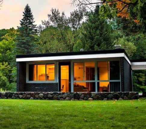 A modern single-story house with large windows and dark brick exterior sits amid lush green grass and tall trees at sunset. Warm lights glow inside, creating a cozy atmosphere.