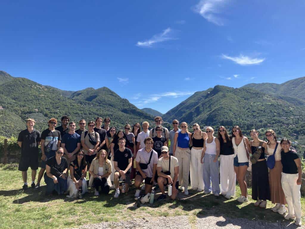 A large group of people poses together outdoors on grass with green mountains and a bright blue sky in the background. Most are smiling and dressed in summer clothing.
