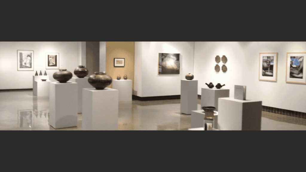 Art gallery interior featuring pottery displayed on white pedestals and framed paintings on the walls. The floor is shiny and the space is well-lit, giving a clean and modern atmosphere.