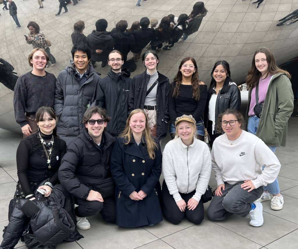 A group of twelve young adults pose and smile together in front of the reflective “Cloud Gate” sculpture, also known as “The Bean,” in Chicago. Their reflections and other people are visible on the shiny surface behind them.