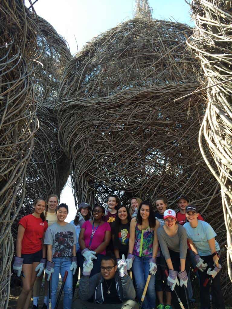 A group of thirteen people wearing gloves and casual clothes poses in front of large, woven stick sculptures outdoors, smiling at the camera. Some hold gardening tools and the weather is sunny and bright.