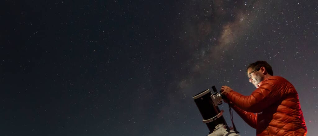 A man in a red jacket is looking through a telescope under a starry night sky. The Milky Way is visible in the background, providing a stunning celestial backdrop to the scene.