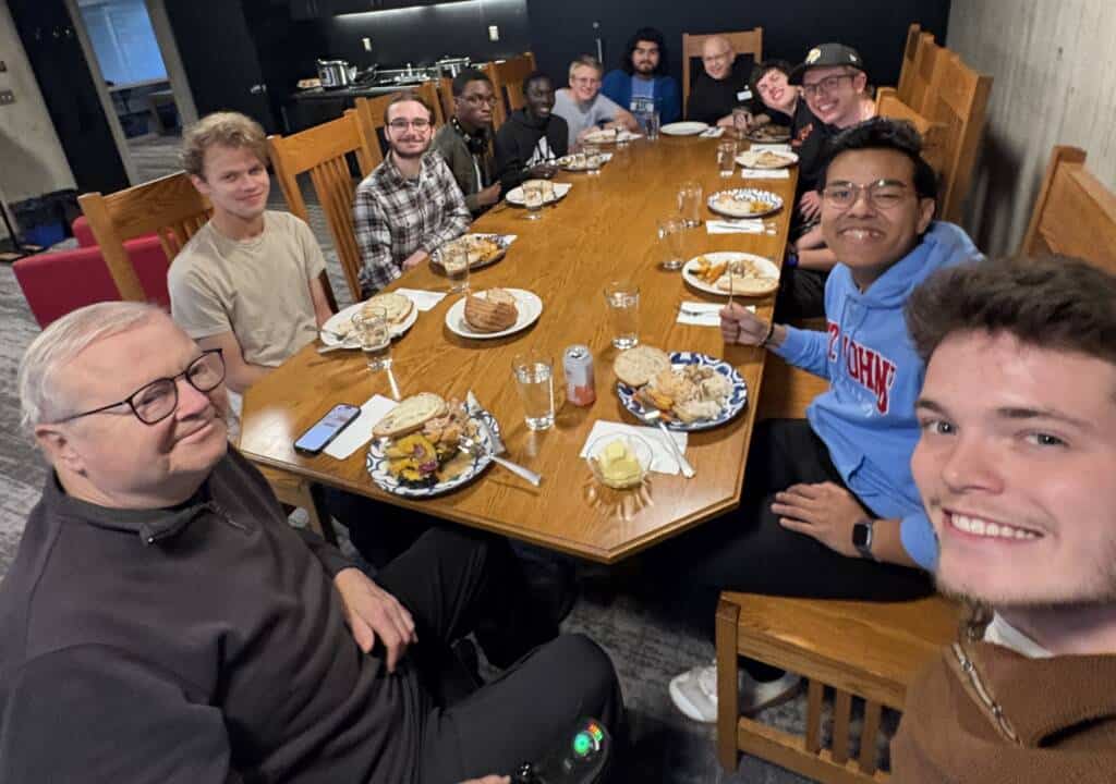 A group of eleven people sit around a wooden dining table sharing a meal, smiling at the camera. The table is set with plates of food, drinks, and bread. The atmosphere appears friendly and casual.