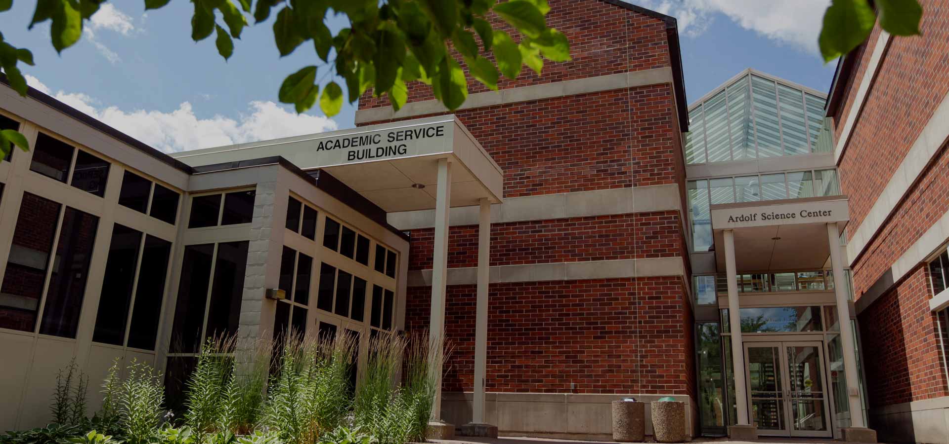 A building complex with red brick walls featuring signs for the "Academic Service Building" and the "Arbolt Science Center." The entrance is framed by white pillars, with tall, green plants in the foreground and tree branches partially obscuring the top.
