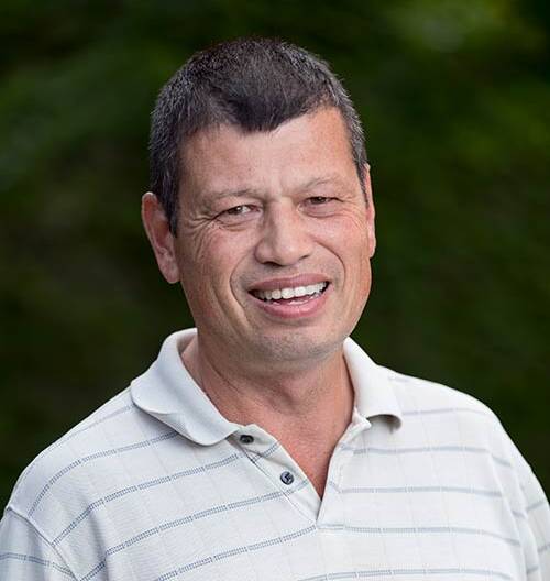 A middle-aged man with short dark hair smiles at the camera. He is wearing a light-colored, short-sleeved, collared shirt with thin horizontal stripes. The background is a blurred natural green setting.