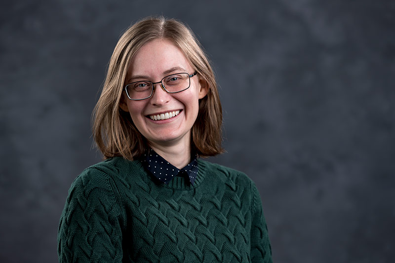 A young person with shoulder-length light brown hair, wearing glasses, a green cable-knit sweater, and a black shirt with white polka dots underneath, smiles at the camera against a gray, textured background.