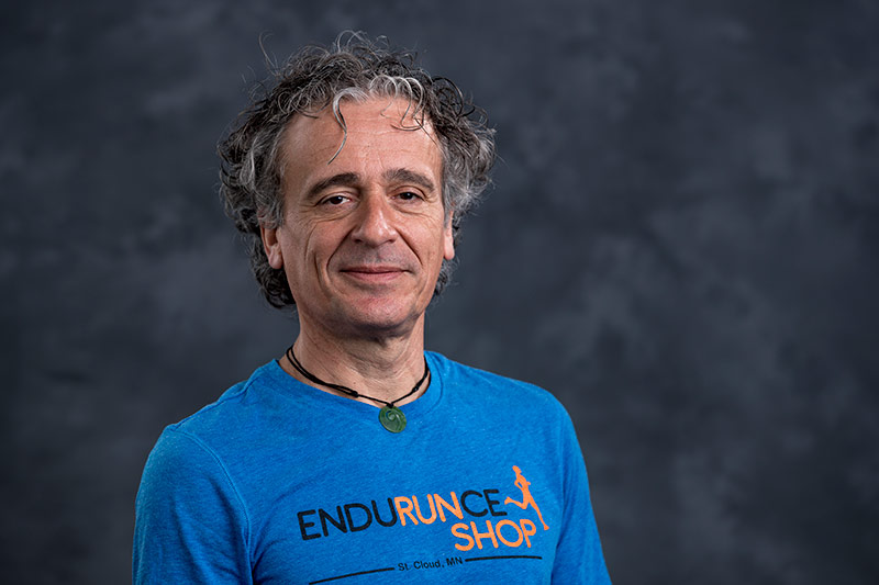 A man with curly gray hair and a slight smile is wearing a blue "Endurance Shop" T-shirt. The background is dark and slightly blurred, putting emphasis on the man.