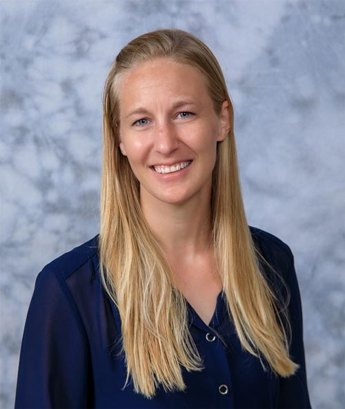 A woman with long, straight blonde hair is smiling in front of a gray marbled background. She is wearing a dark blue blouse.