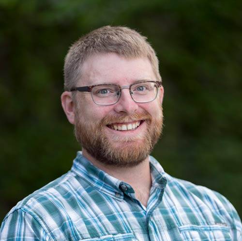 A man with short light brown hair and a beard smiles at the camera. He is wearing glasses and a green and white plaid shirt. The background is out of focus greenery.