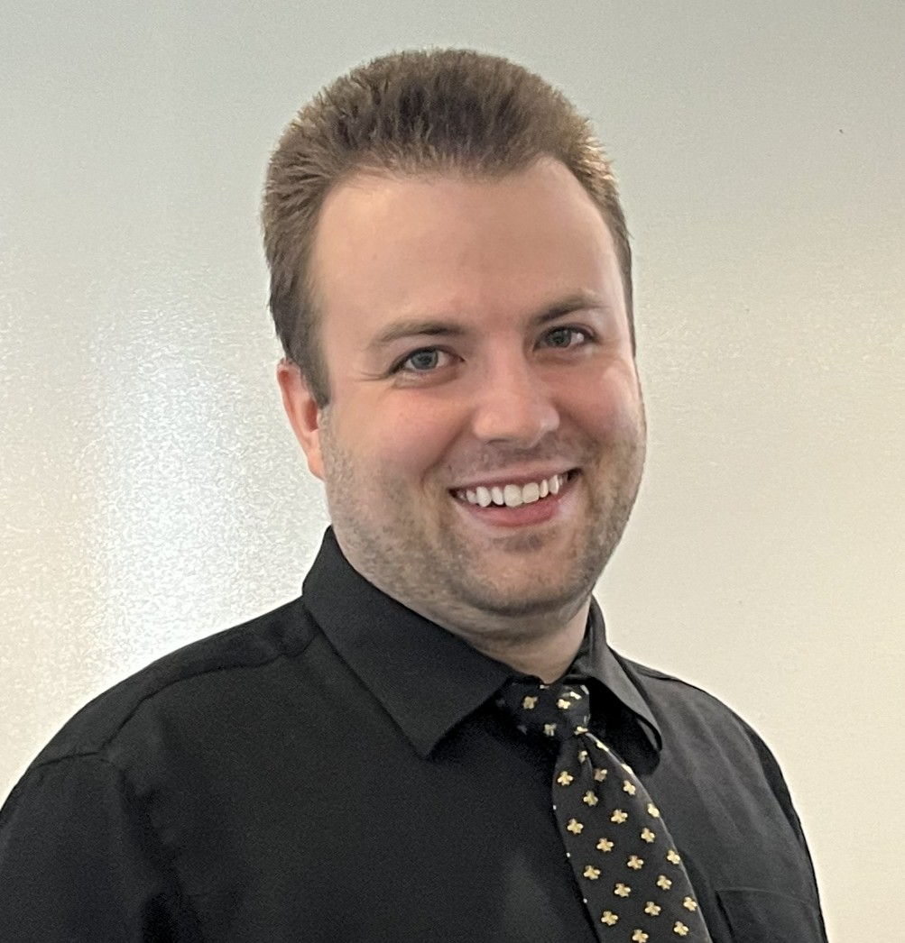 A smiling man with short brown hair wearing a black dress shirt and a black tie with a gold pattern, standing in front of a plain light-colored background.