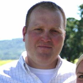 A man with short hair is standing outside on a sunny day, wearing a white collared shirt with a faint checkered pattern. There is greenery and a blurred landscape in the background.