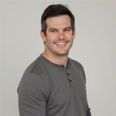 A man with short dark hair and light stubble smiles at the camera. He is wearing a long-sleeve, gray, henley shirt and is posed against a plain light gray background.