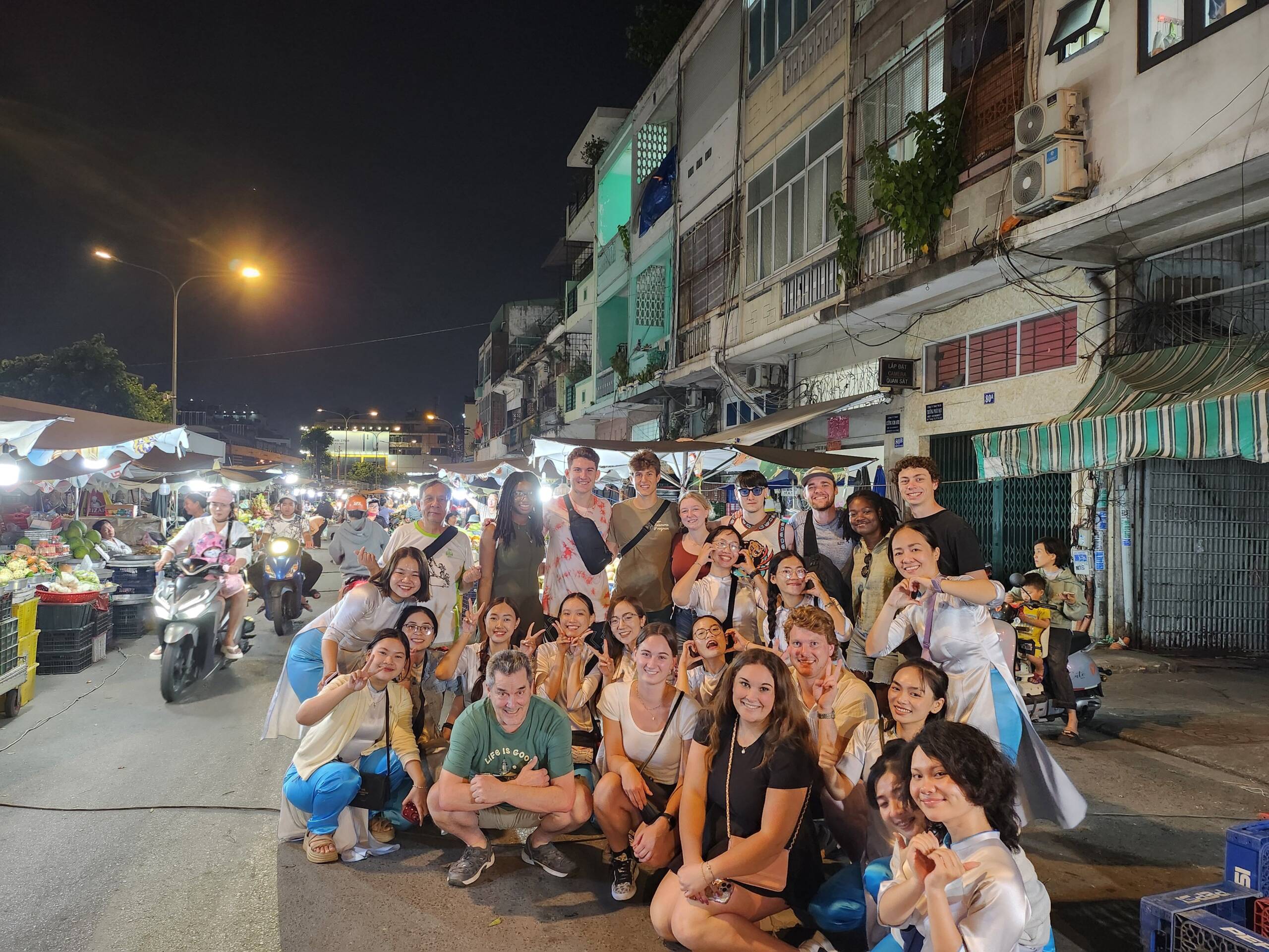 A large group of people, some in costumes, pose and smile for a photo at night in a lively street market with stalls, shops, and scooters in the background.