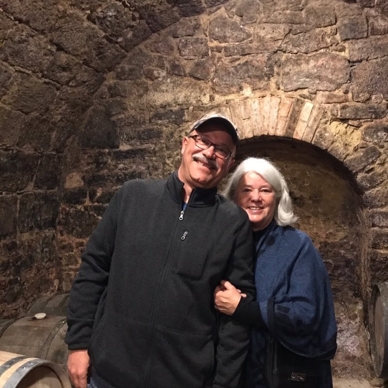 A smiling older man and woman stand close together in a stone-walled cellar, with arched brickwork behind them and a wooden barrel nearby. The man wears glasses and a cap; the woman has gray hair and a dark jacket.
