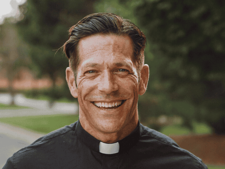 A smiling man wearing a black clerical shirt with a white collar stands outdoors with green trees and grass in the background.