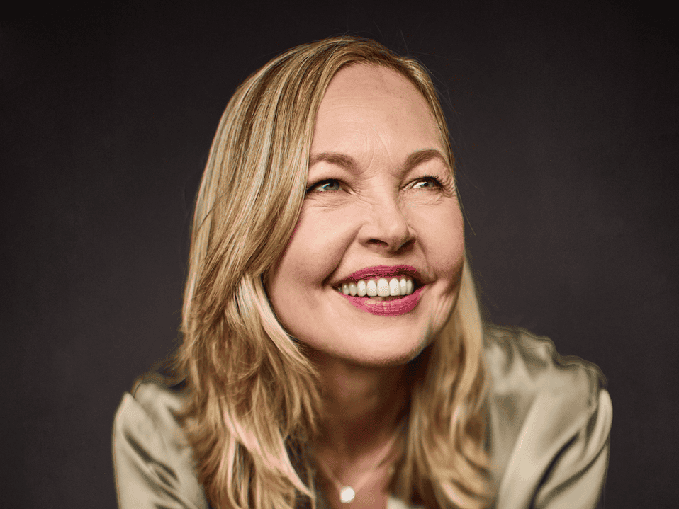 A woman with blonde hair wearing a shiny, light-colored blouse and jeans sits against a dark background, smiling brightly and leaning forward with her hands clasped together.