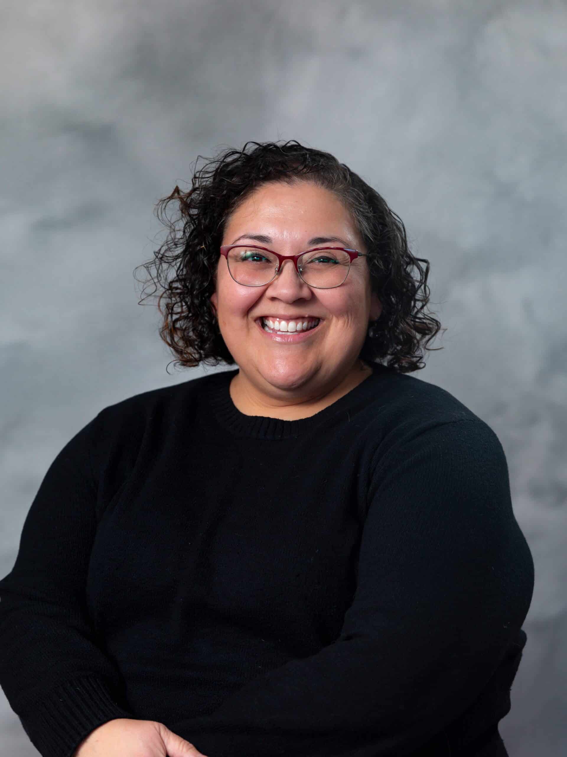 A woman with curly hair and glasses smiles warmly at the camera. She is wearing a black sweater and sits against a neutral gray background.