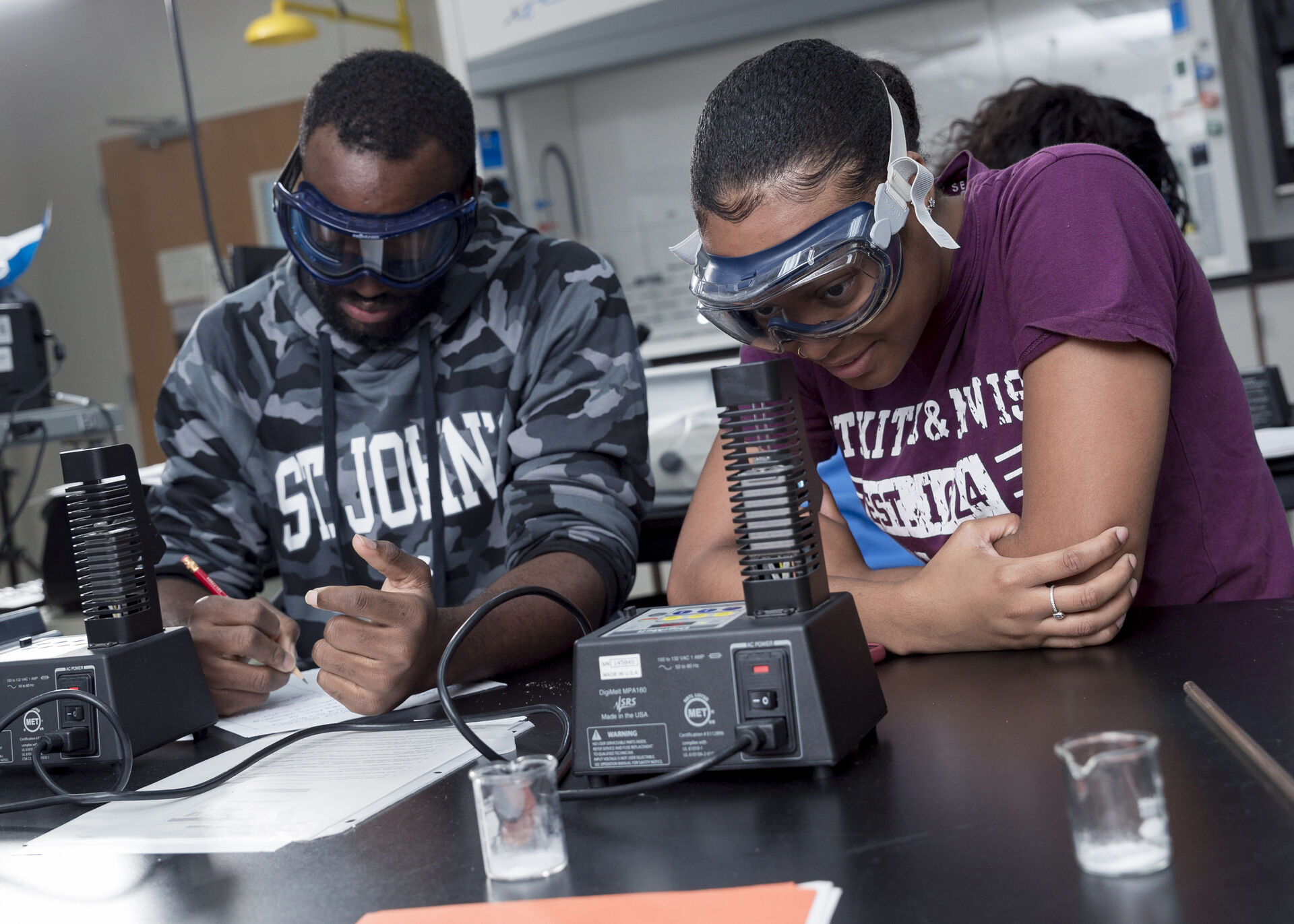 Two students wearing safety goggles work at a lab station with papers and beakers. One student takes notes while the other seems focused on a task. They are in a classroom setting with lab equipment visible in the background.