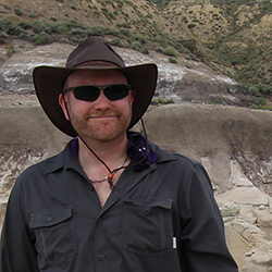 A man with a beard, wearing dark sunglasses and a wide-brimmed hat, is standing in a rugged outdoor area with rocky hills in the background. He is dressed in a dark, long-sleeved shirt and has a slight smile on his face.