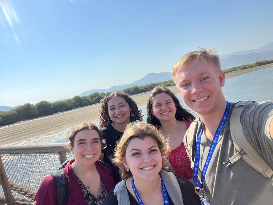 Five smiling young adults posing for a selfie outdoors by a lake, with mountains and a clear blue sky in the background. Some are wearing lanyards and backpacks, and sunlight brightens the scene.