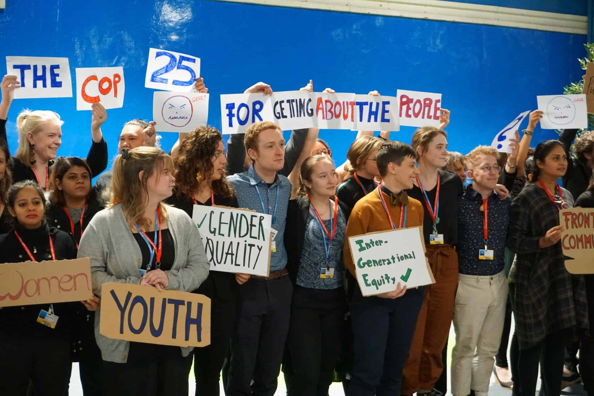 A diverse group of people stands together holding signs with messages like "Gender Equality," "Youth," and "COP 25 for the People," advocating for equity at a public event or conference.