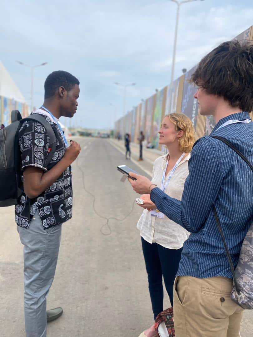 Three people stand on a wide pathway outdoors, two holding notepads and a recorder, interviewing a man with a backpack. They appear focused, engaged in conversation under a cloudy sky.