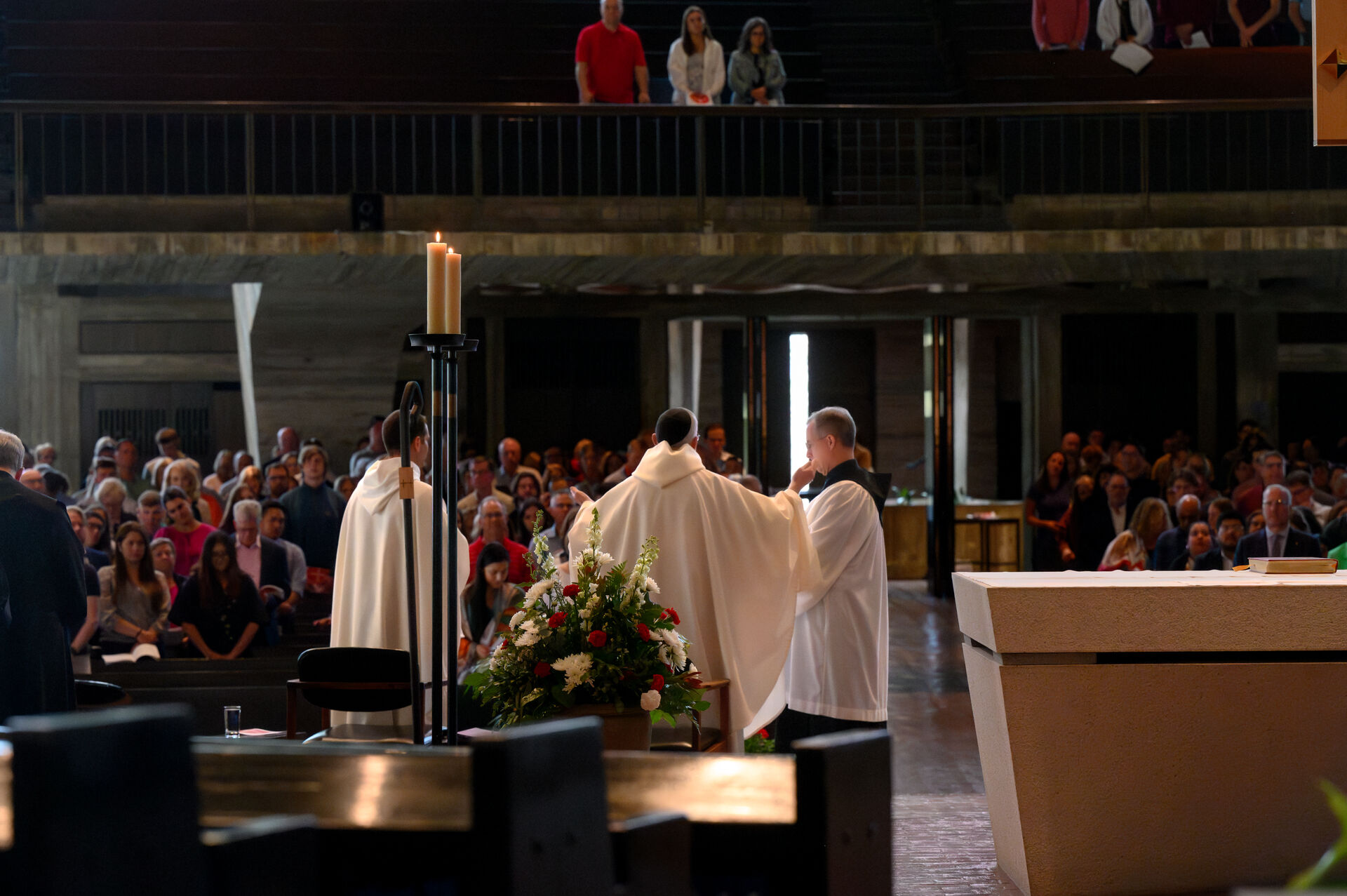 Three clergy members in white robes stand at an altar during a church service. One holds a microphone. A congregation is seated in pews, watching the ceremony. Flowers adorn the altar.