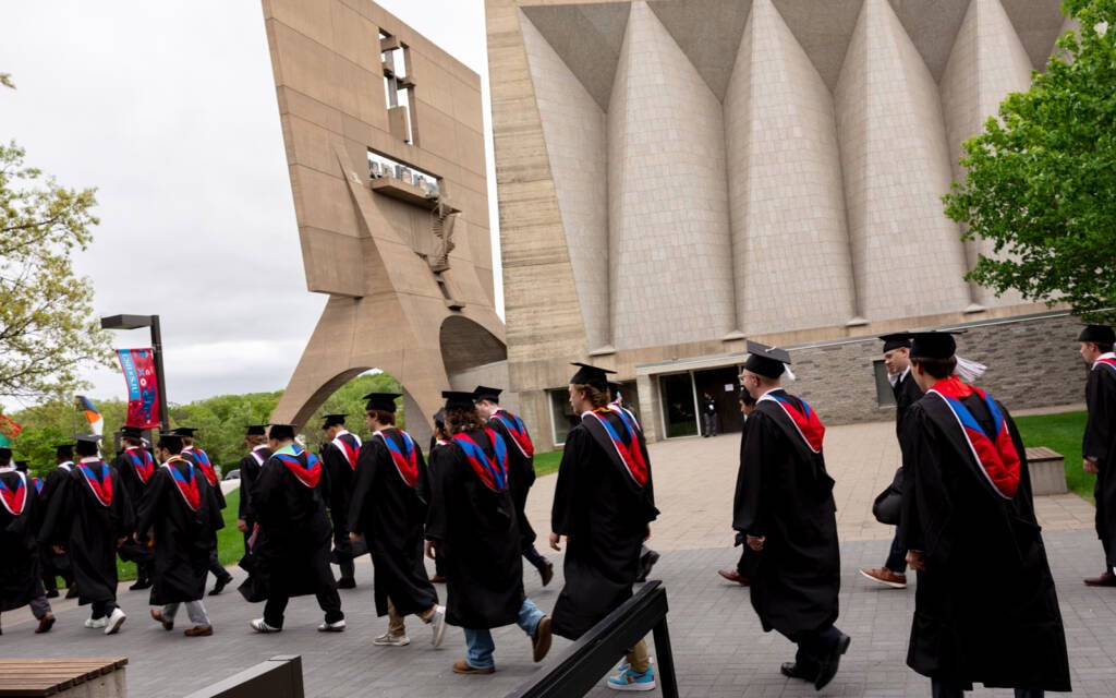 A group of graduates in caps and gowns walk together outside a modern campus building with geometric architecture and a large concrete bell tower. The scene is lively, with greenery and trees in the background.