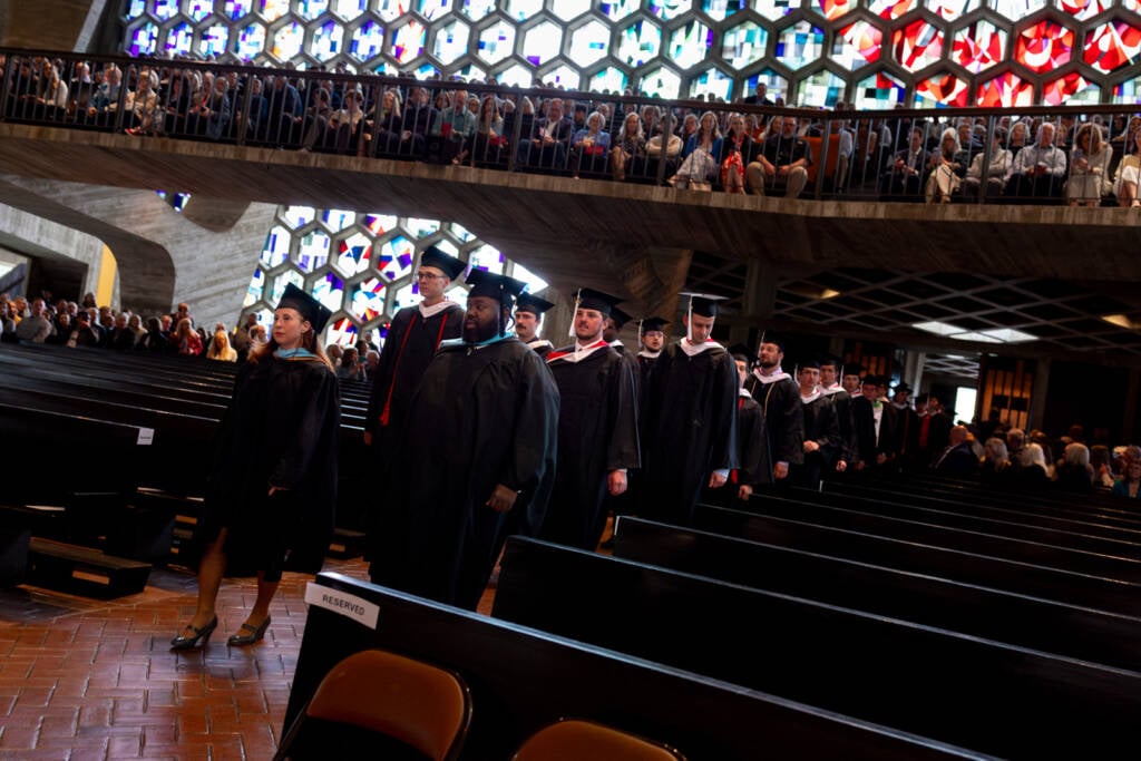 Graduates in caps and gowns walk down the aisle of a church filled with seated guests; colorful stained glass windows light the space and an upper balcony holds more people watching the ceremony.