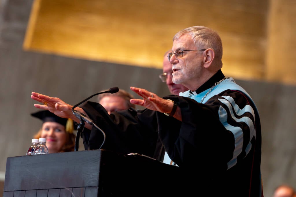An older man in academic regalia speaks at a podium, gesturing with both hands outstretched. A woman in a graduation cap and gown smiles in the background. The setting appears to be a graduation ceremony.