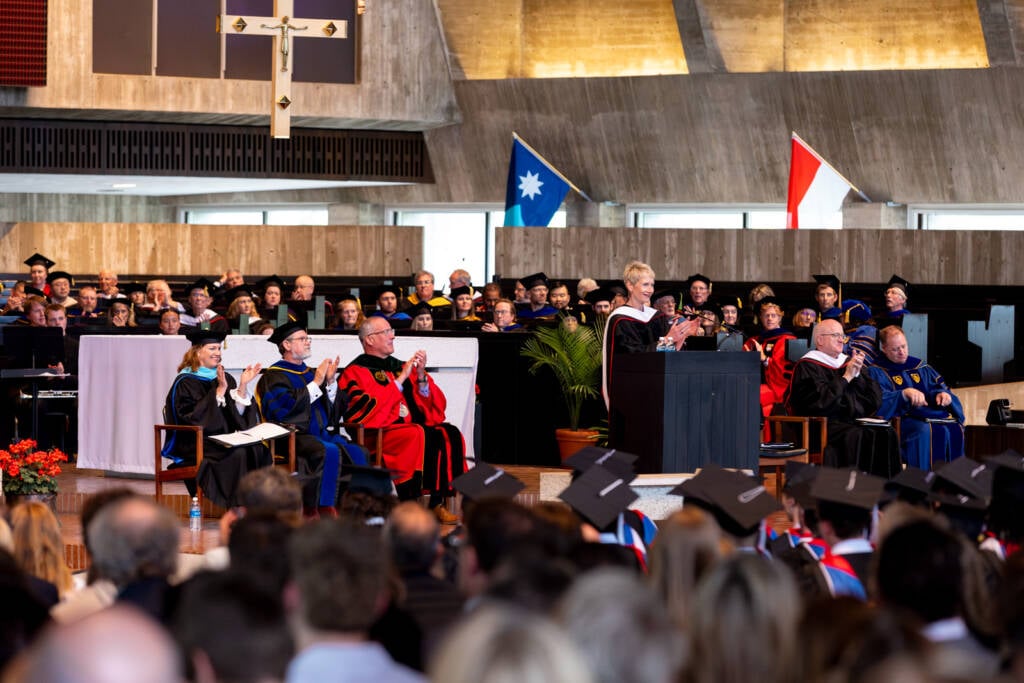 A woman in academic regalia speaks at a podium on stage during a graduation ceremony, with faculty members seated behind her and an audience of graduates in caps and gowns in the foreground.