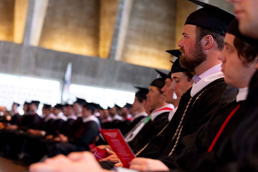 Rows of graduates in caps and gowns sit attentively during a ceremony, holding programs. The background is softly lit, highlighting the celebratory atmosphere.