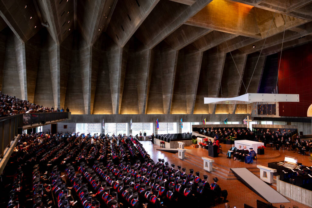 Graduates in caps and gowns sit in rows inside a modern, spacious church with high concrete ceilings during a commencement ceremony, while an audience watches from the side. The altar and clergy are visible at the front.
