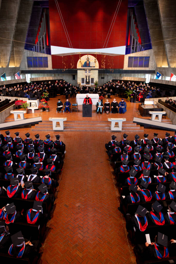 A large group of graduates in caps and gowns sit in rows facing a stage inside a modern church. Faculty in academic regalia are seated on stage, and a speaker stands at a podium. The church has high ceilings and stained glass windows.