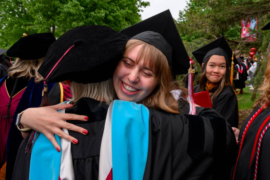 A graduate in a cap and gown joyfully hugs another person in academic attire at an outdoor graduation ceremony, while another graduate smiles in the background. Trees and other graduates are visible.