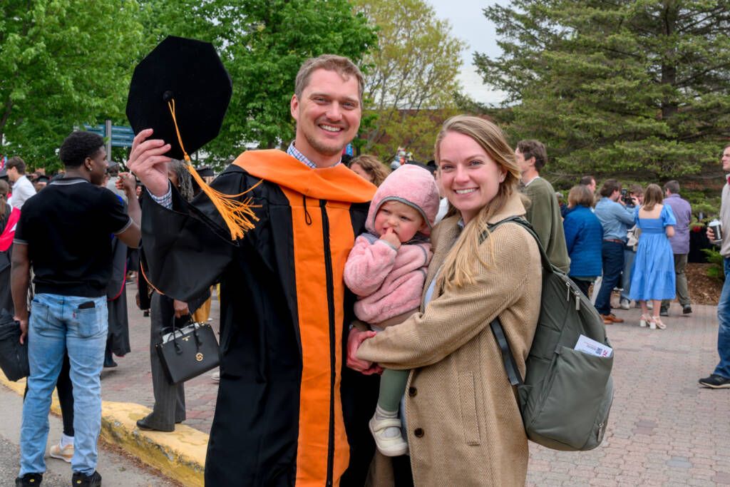 A smiling graduate in a cap and gown stands outdoors with a woman holding a baby. The woman and baby are both smiling. Other people are gathered in the background among trees.