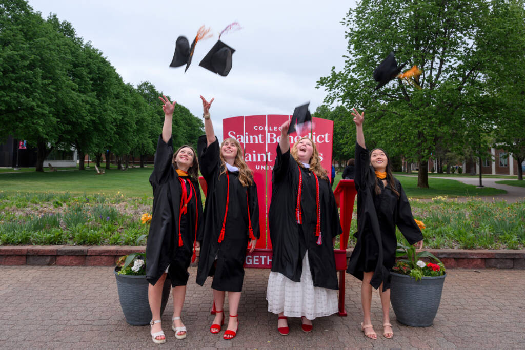 Four graduates in black gowns and caps joyfully throw their caps in the air in front of a College of Saint Benedict sign, celebrating outdoors with green trees and flowers in the background.