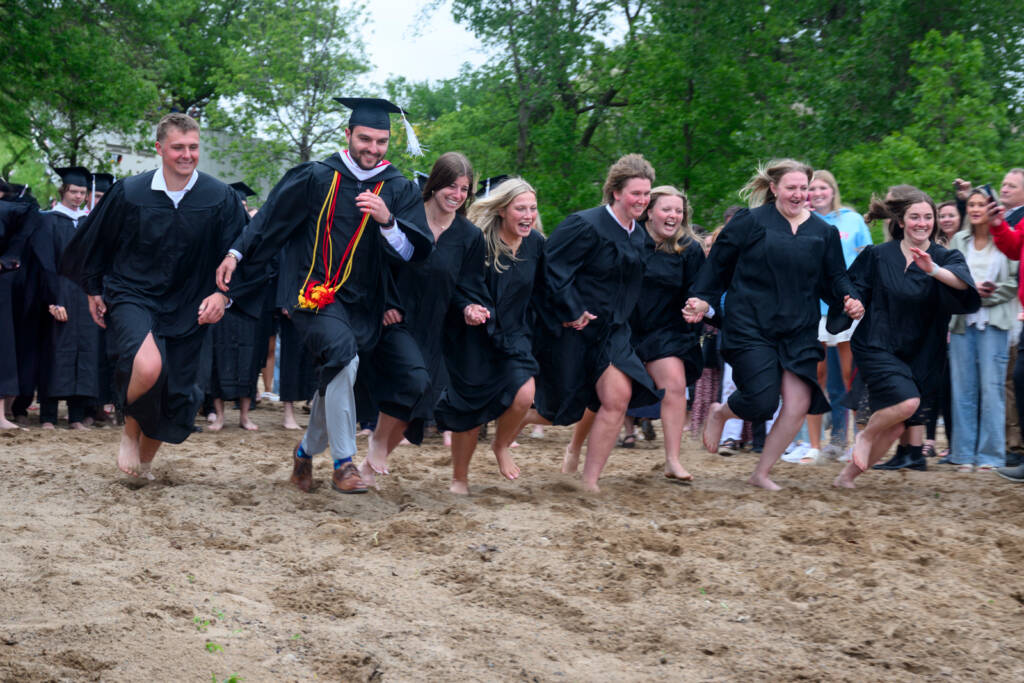 A group of graduates in black gowns, some barefoot, laugh and run together across a sandy area outdoors, while a crowd of people watches in the background among green trees.