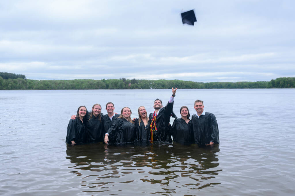 Eight graduates wearing black gowns stand waist-deep in a lake, smiling at the camera. One person is tossing a graduation cap into the air. Trees and a cloudy sky are in the background.