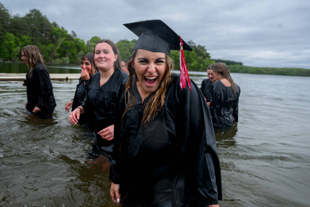 A group of graduates in black caps and gowns stand in a lake, smiling and laughing. The main focus is on a woman in the foreground who is grinning joyfully at the camera. Trees and cloudy sky are visible in the background.