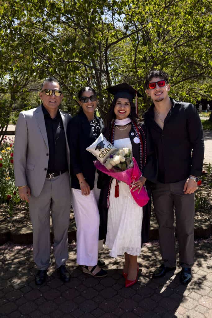 A young woman in a graduation cap and gown holds flowers and smiles, standing outdoors with three people, likely family, all dressed formally. Trees and plants are in the background on a sunny day.