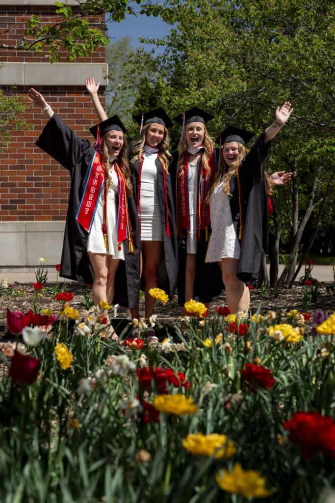 Four young women in graduation caps and gowns stand outdoors by blooming flowers, smiling and raising their arms in celebration. Trees and a brick building are in the background, and it is sunny.
