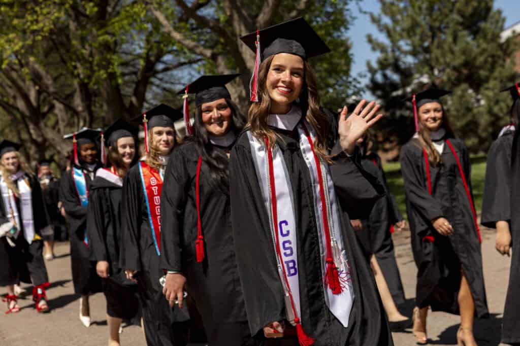 A group of graduates in caps and gowns walk outdoors in a line. One woman in front smiles and waves at the camera; others follow behind her. Trees and a sunny sky are visible in the background.