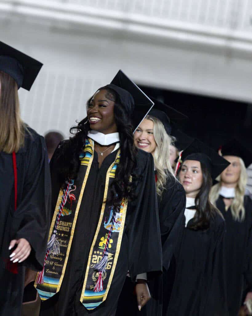 A group of graduates in caps and gowns smile as they walk in a graduation ceremony; the woman in front wears a colorful stole and looks to the side.