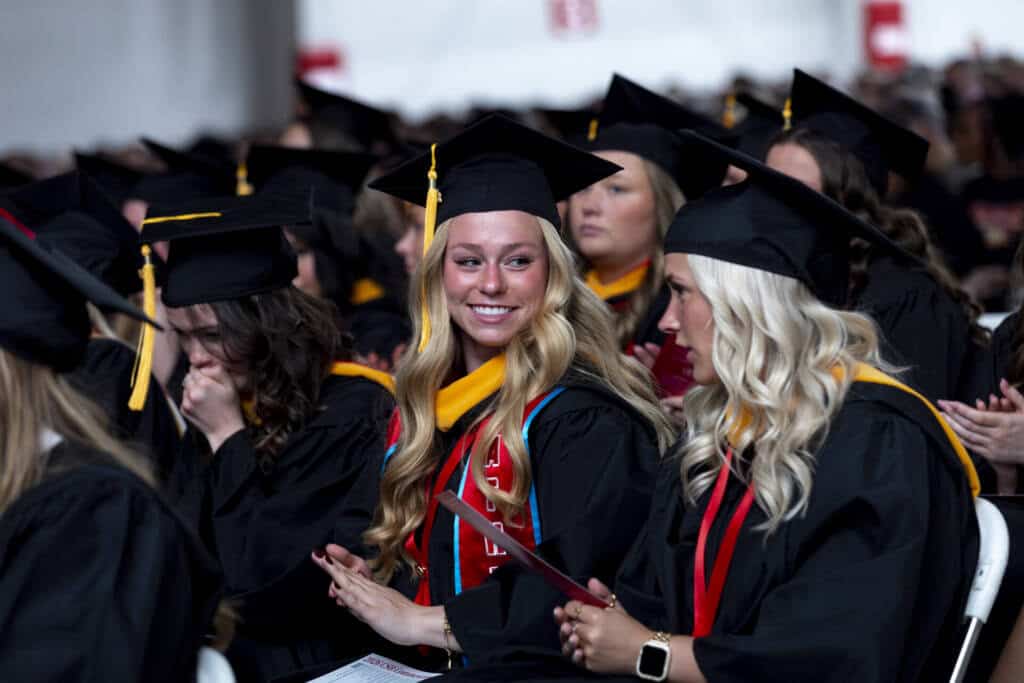 A group of graduates in caps and gowns sit during a ceremony. One woman with long blonde hair smiles at a woman beside her, while others sit and look forward in the background.